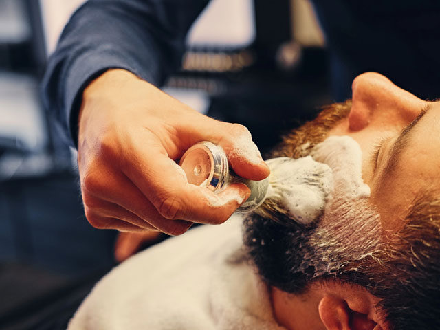 A barber applying the lather of a shaving soap puck mixed with warm water via a shaving brush